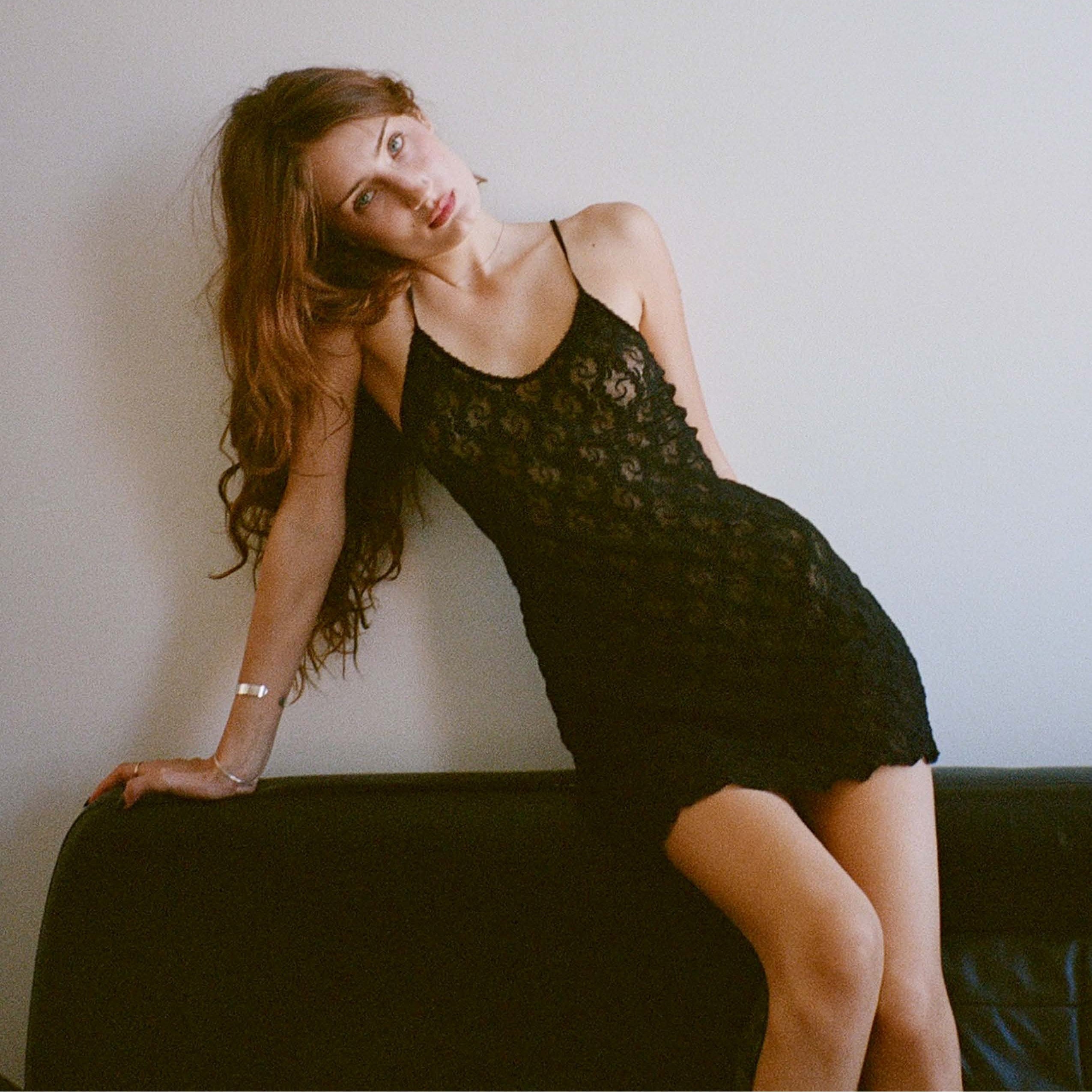Woman in a black lace dress sitting on a couch against a plain wall.